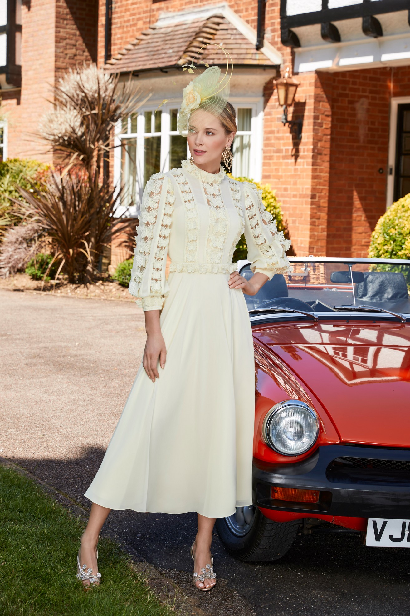 Woman standing in front of a house in lemon yellow special occasion dress with a-line skirt, long sleeves and 3d butterfly detailing with matching fascinator hat 